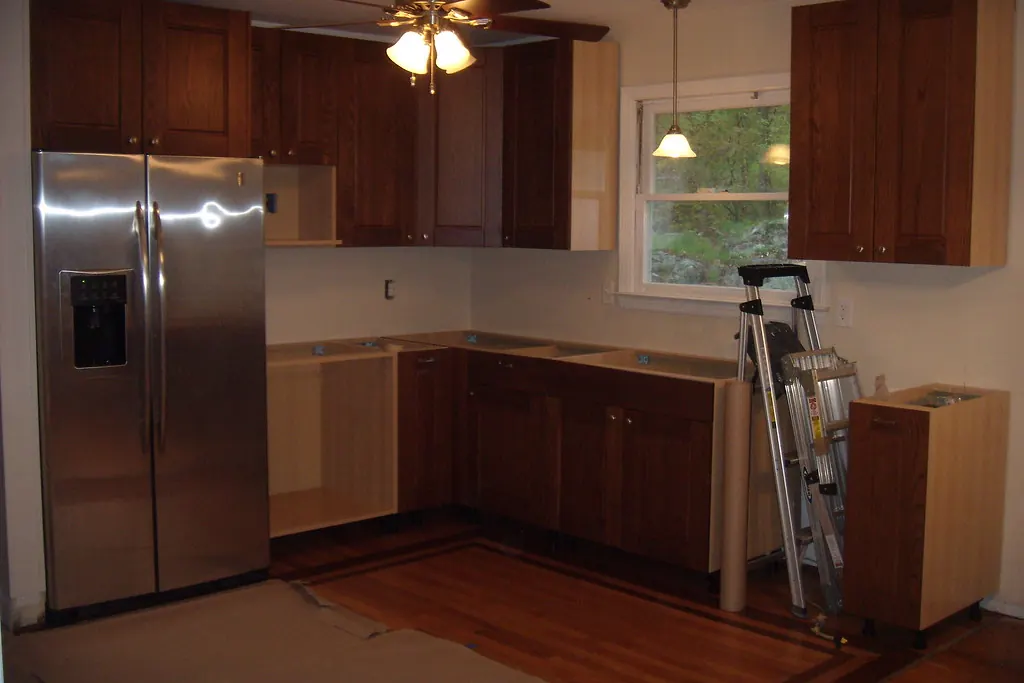A kitchen mid-installation with stained base and wall cabinets in place, a bare floor and a ladder, in a house of the kind found in San Clemente CA