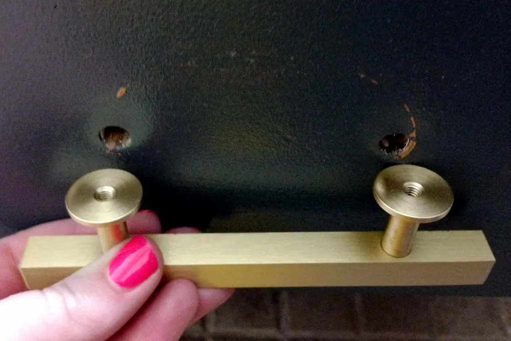 A hand holding a brass bar pull against a dark cabinet door with two drilled holes, during a cabinet hardware change in San Clemente CA
