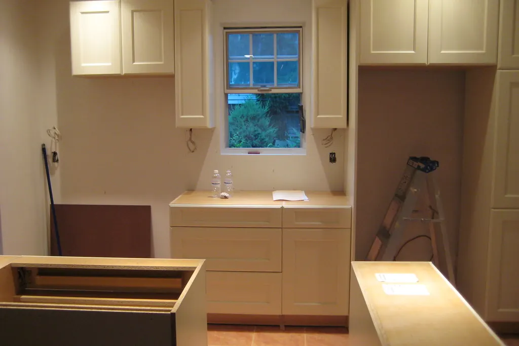 Cream cabinets partly installed with a base cabinet standing loose on the floor and a ladder in the room, during cabinet work in a San Clemente CA house